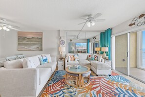 A spacious living room with a large window, a ceiling fan, and a colorful rug.