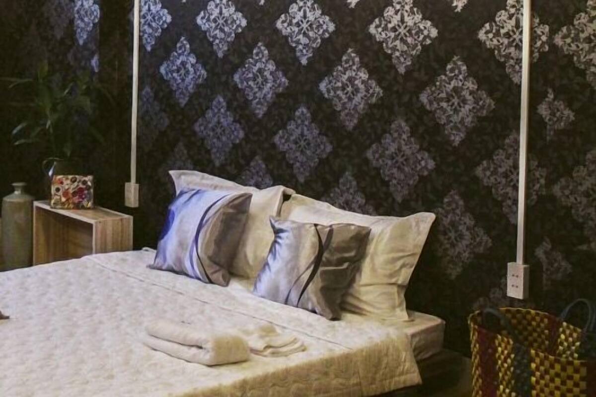 Cama bem arrumada com travesseiros e cobertor, mesa de cabeceira pequena com uma xícara, e uma cadeira com almofada.