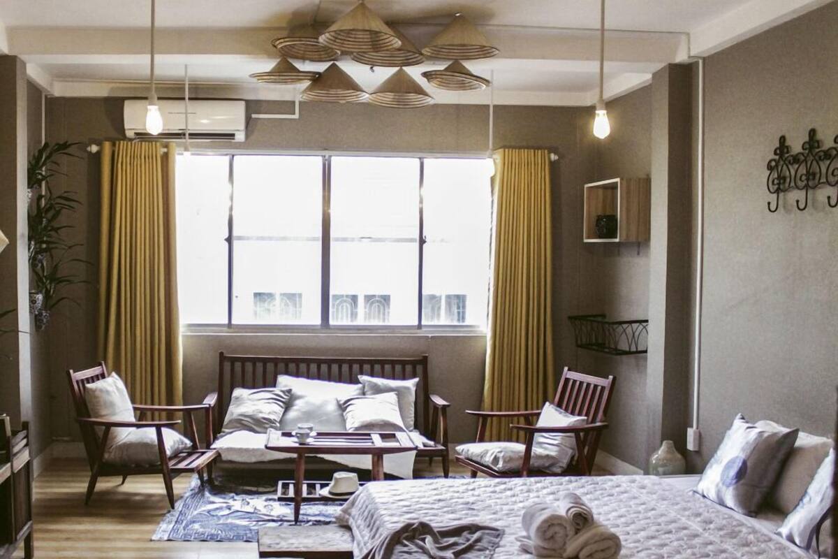 Quarto de hotel com cama, duas cadeiras, uma mesa pequena, janela com cortinas e ar-condicionado fixado na parede.