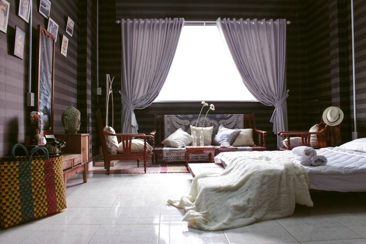 Quarto com cama, duas cadeiras, uma mesinha, janela com cortinas e parede com quadros emoldurados.