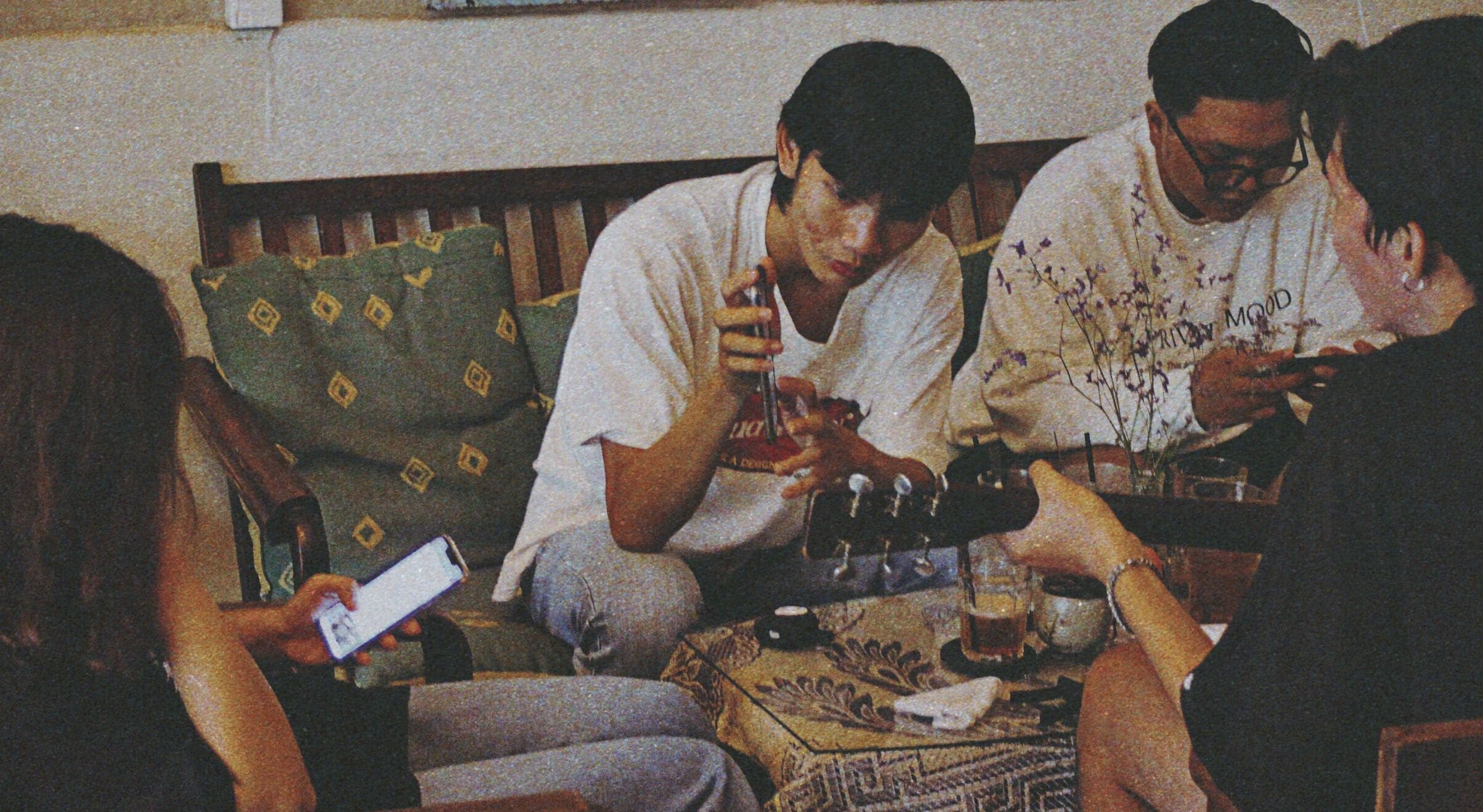 A group of people sitting on a couch, some using a smartphone, with a table in front of them holding drinks and a small decorative item.