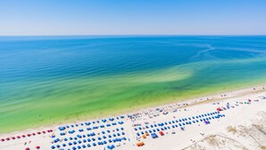 An aerial view of a beach with rows of blue and red beach umbrellas, a sandy shore, and the ocean.