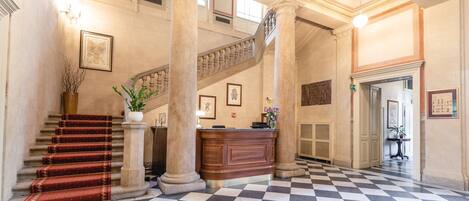 Un vestíbulo con piso a cuadros, una escalera, una mesa de recepción y columnas.