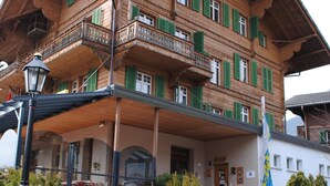 A traditional wooden building with balconies and green shutters, featuring a stone entrance and a flag. - Hotel Post Vista (Zweisimmen)