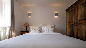 Una cama bien hecha con edredón blanco y almohadas decorativas, un armario de madera, una mesita de noche con lámpara y dos luces fijadas a la pared.