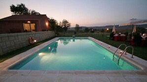 Una piscina rectangular con borde de piedra, rodeada por un muro de piedra y un césped con mesas y sombrillas.