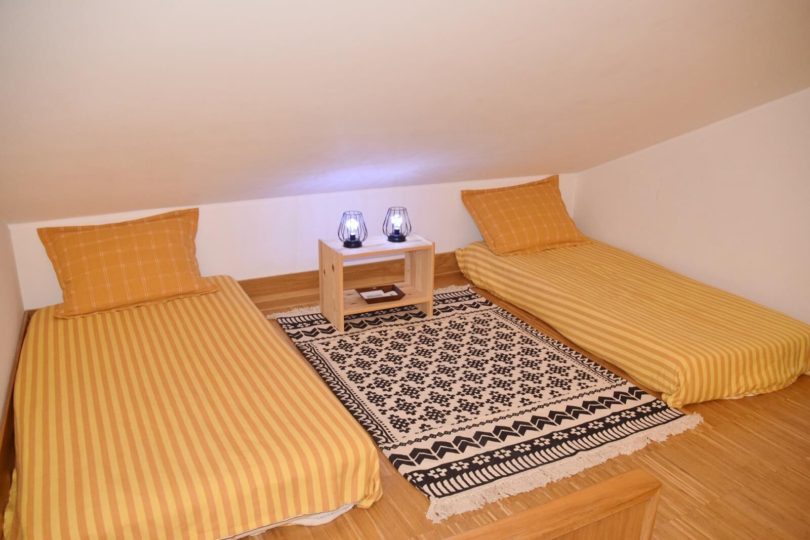 Una habitación pequeña y acogedora con dos camas individuales, una mesita de noche de madera con dos lámparas y una alfombra estampada.