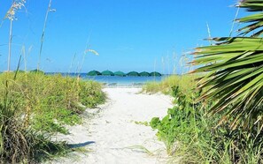 A sandy beach path leading to a calm sea with green umbrellas in the distance.