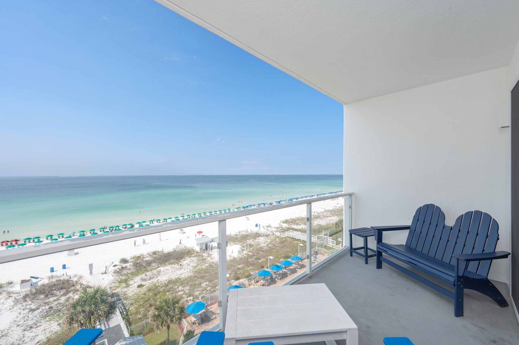A balcony with a view of the beach, a table, and two Adirondack chairs.