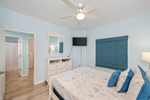 Een slaapkamer met een bed, een plafondventilator, een televisie en een raam met gordijnen.