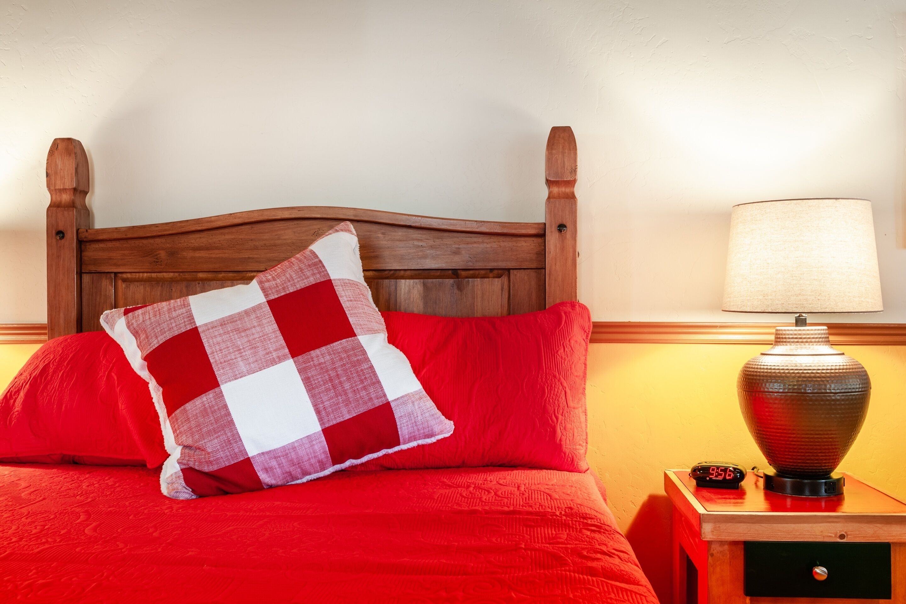 A bed with a wooden headboard, red bedding, a checkered pillow, a bedside lamp, and a digital clock.