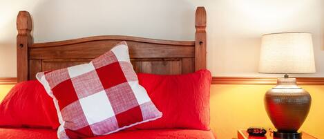 A bed with a wooden headboard, red bedding, a checkered pillow, a bedside lamp, and a digital clock.
