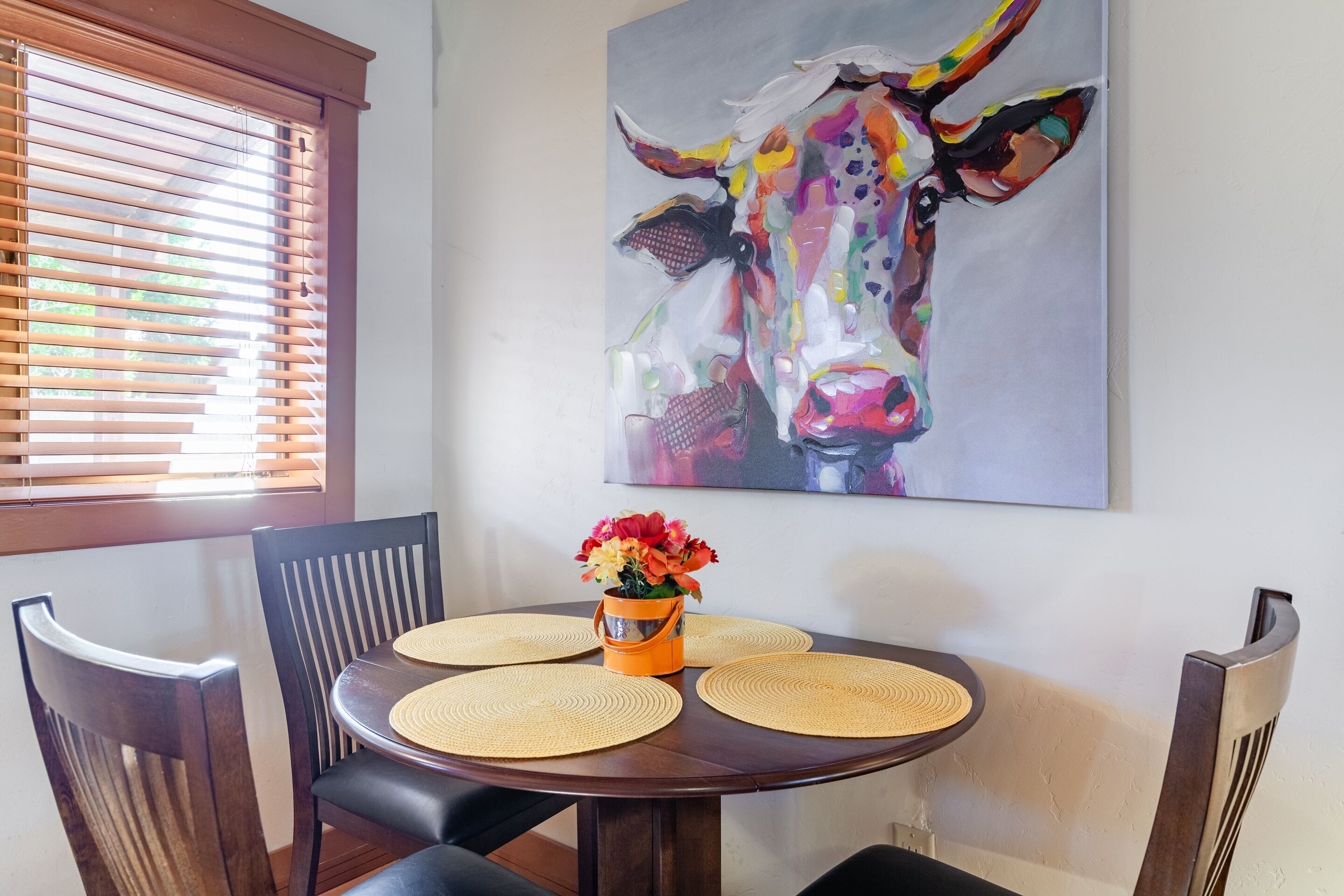A dining area with a round table, four chairs, a vase with flowers, and a colorful cow painting on the wall.