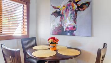 A dining area with a round table, four chairs, a vase with flowers, and a colorful cow painting on the wall.