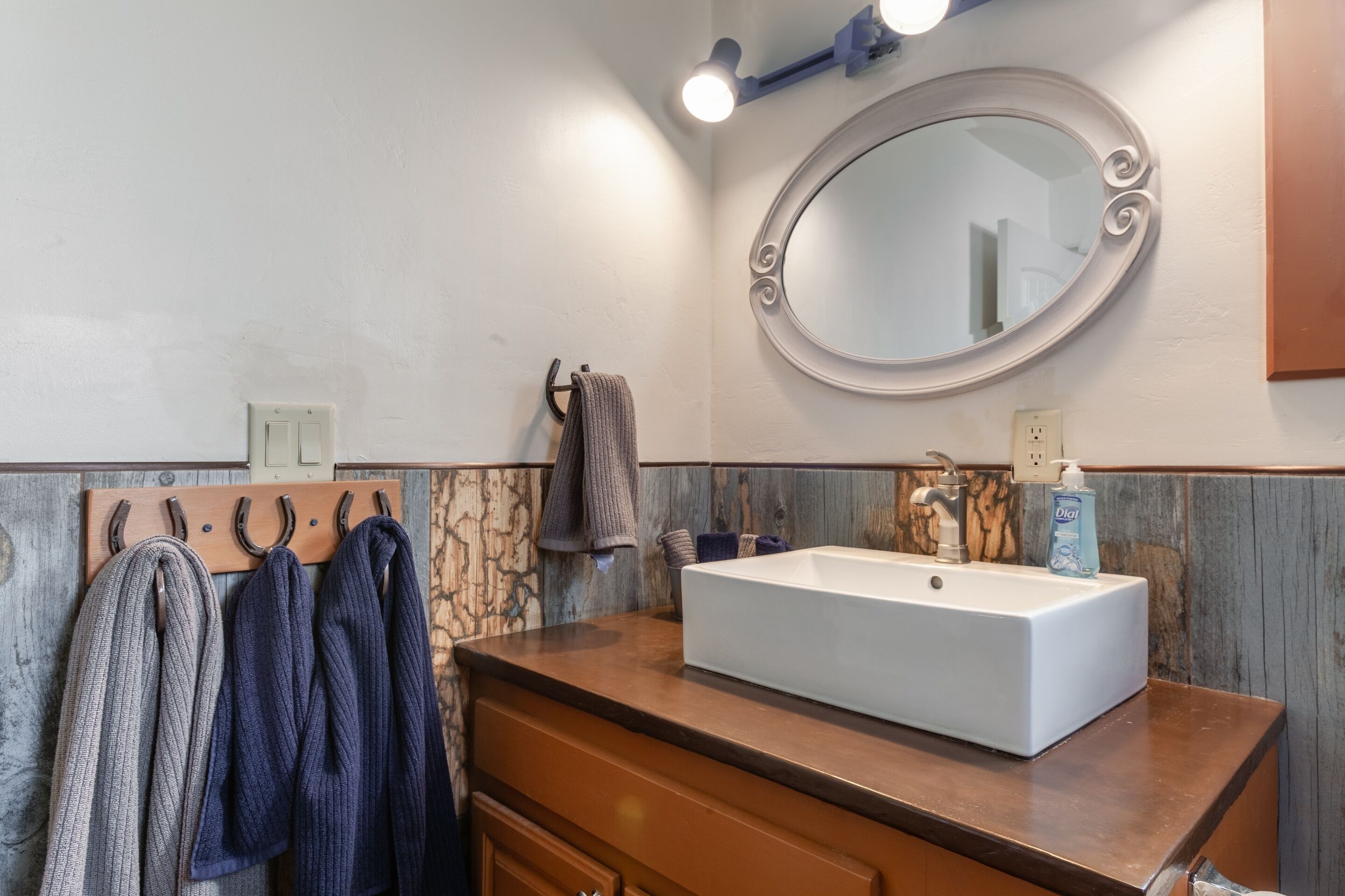Un baño con lavamanos blanco, tocador de madera, toallero y espejo con marco decorativo.