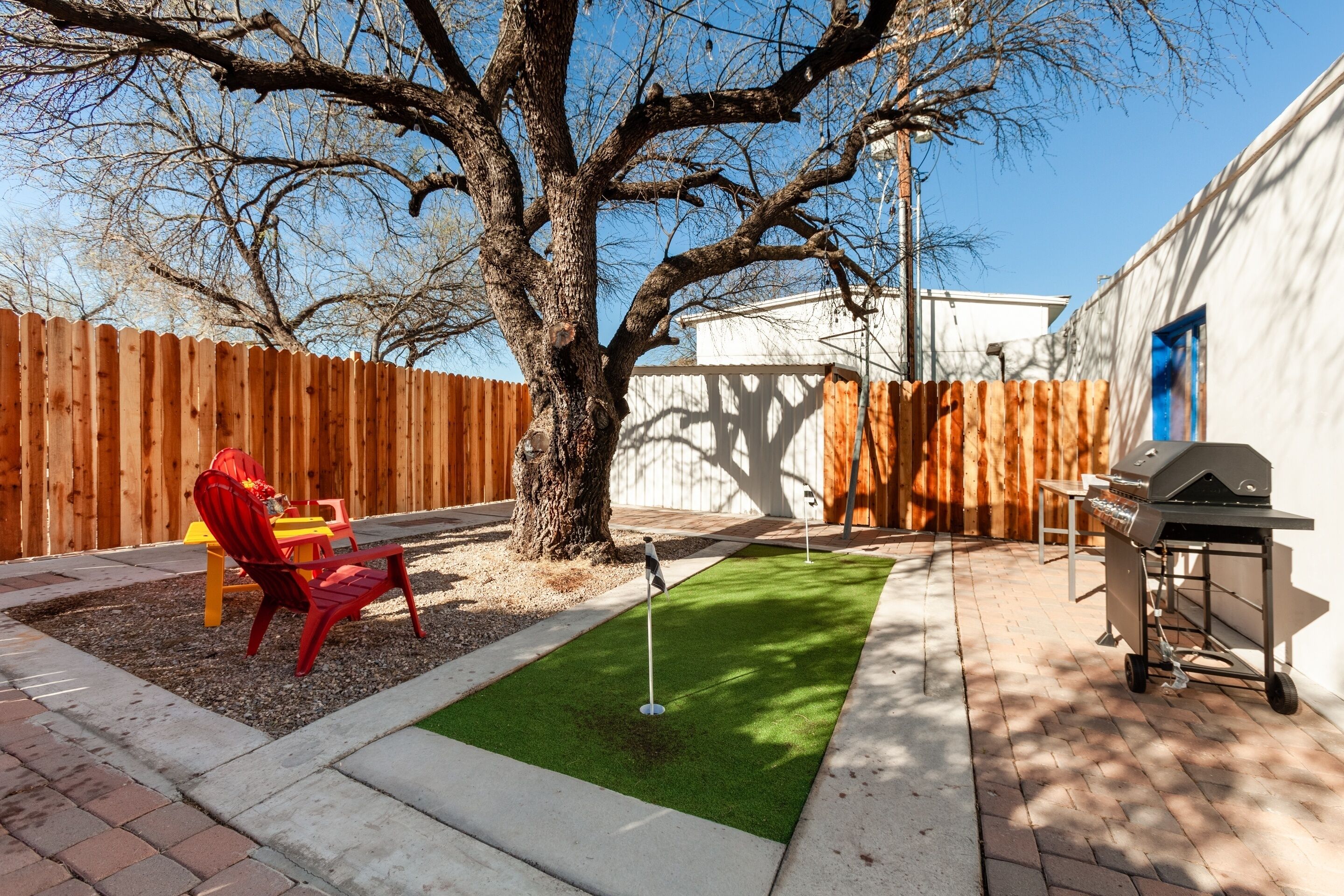 A backyard with a wooden fence, a tree, a red chair, a grill, and a green artificial turf area.