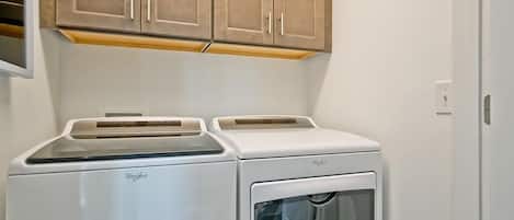 A compact laundry room with a front-loading washer and dryer, wooden cabinets above, and a white door to the right.