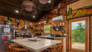 Cucina di una baita in legno con isola centrale, sedie di legno e un'ampia finestra vista su del verde.