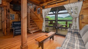 Interno di una baita in legno con un divano, una panca e una scala che conduce a un balcone con vista.