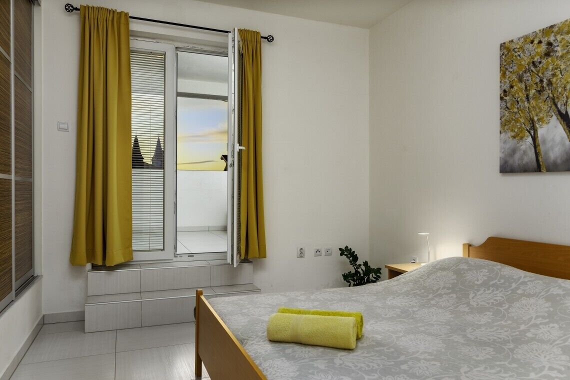 A bedroom with a bed, a window with yellow curtains, a nightstand, and a wall-mounted painting.
