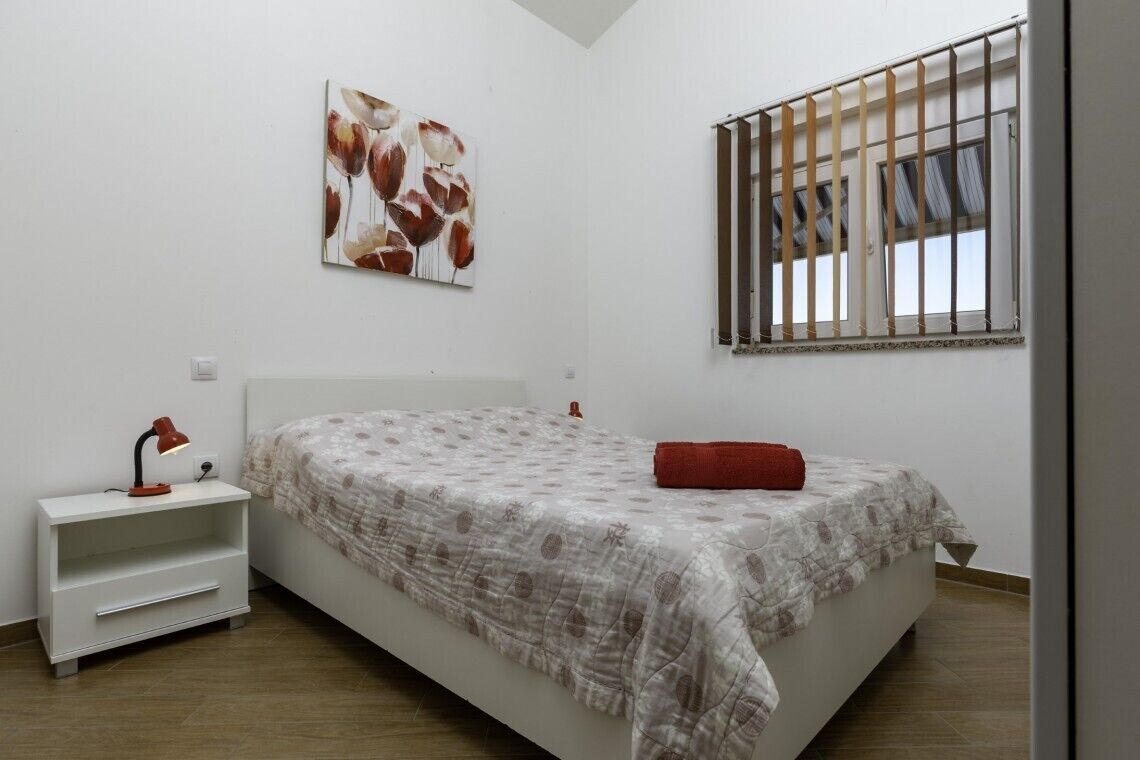 Une chambre à coucher avec un lit, une table de chevet, une lampe, une fenêtre avec des barreaux et une œuvre d’art murale.