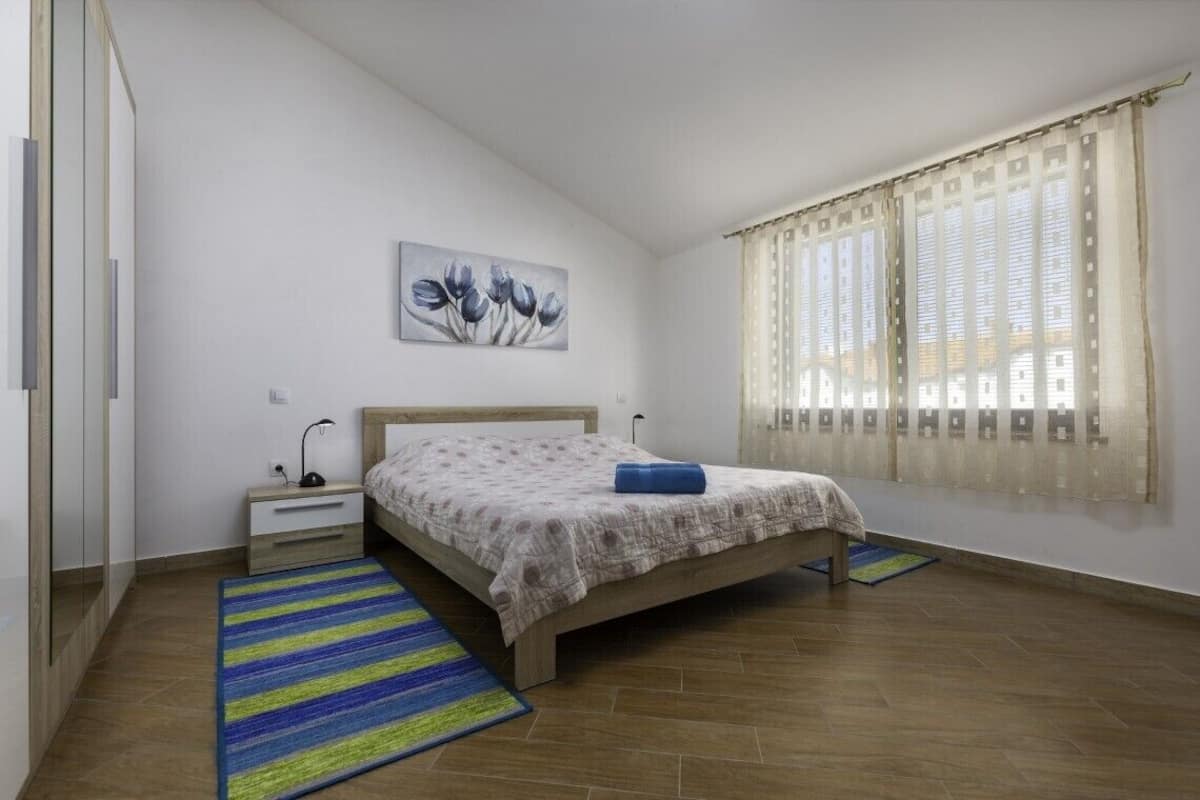 Un dormitorio con cama, mesita de noche, lámpara, un cuadro y ventana con cortinas.