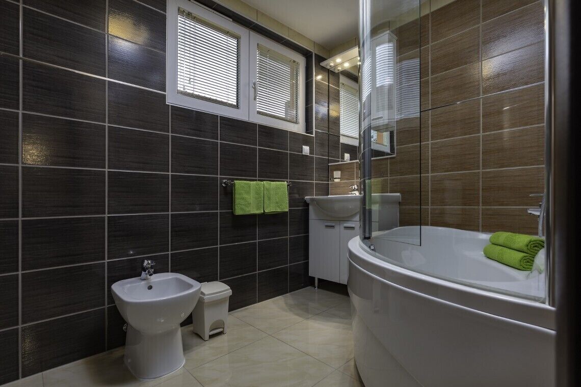 Un bagno moderno con pareti piastrellate in tonalità scura, un lavandino bianco, un bidet, un water e una vasca da bagno ampia.
