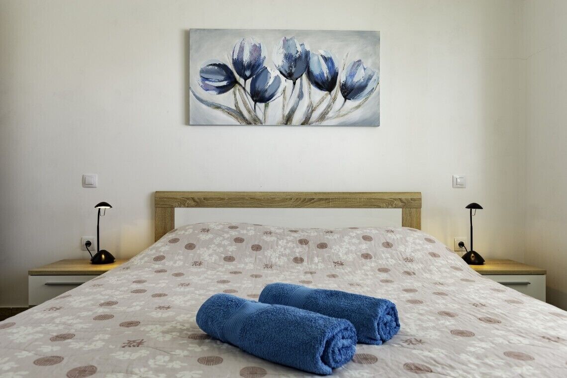 A bedroom with a bed, a painting of flowers, two bedside lamps, and a wooden headboard.