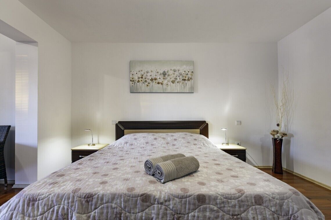 A neatly made bed with a patterned bedspread, two bedside lamps, a wall-mounted artwork, and a vase with dried plants.