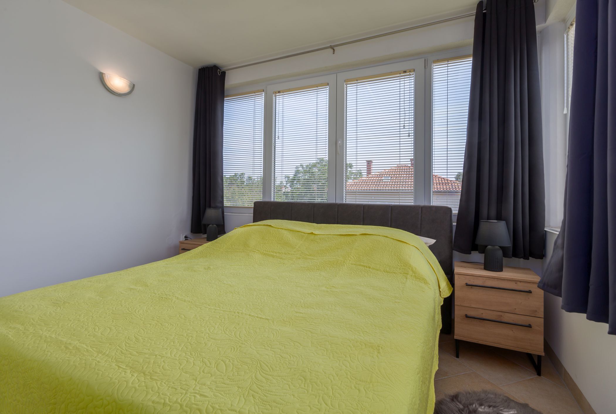Een slaapkamer met een bed, nachtkastjes, een raam met zonwering en uitzicht op gebouwen.