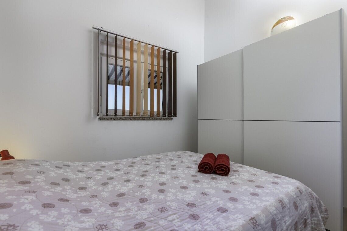 A bedroom with a bed, a window with blinds, a wardrobe, and a wall-mounted light fixture.