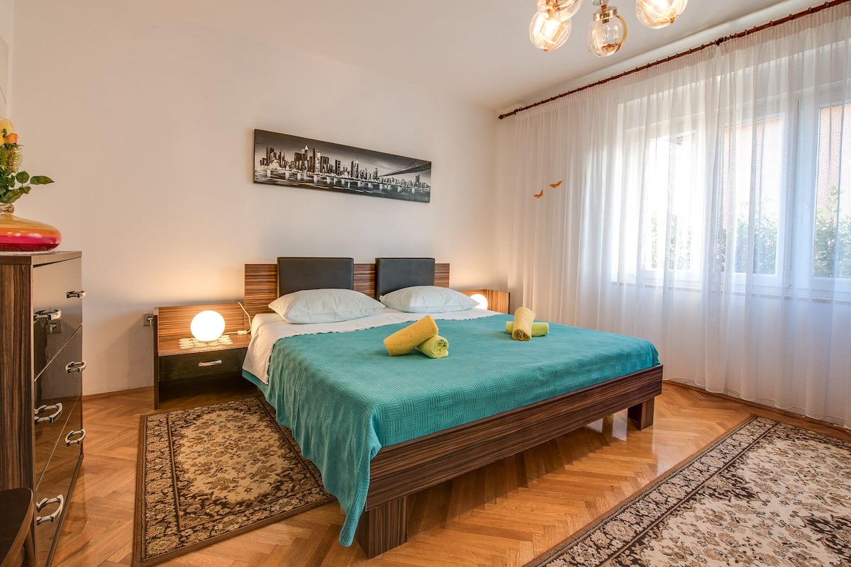 Ein Schlafzimmer mit Bett, Nachttischen, einem Fenster mit durchsichtigen Vorhängen und einem Stadtbild.