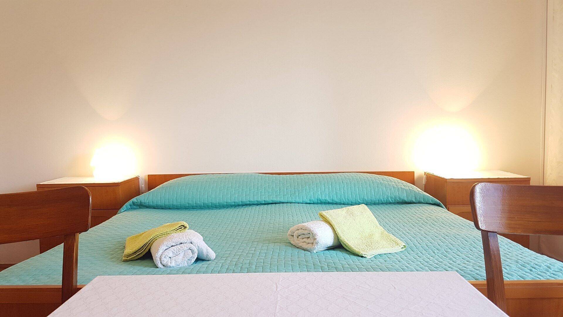 A single bed with a teal quilt, two rolled towels, and a white table in front.