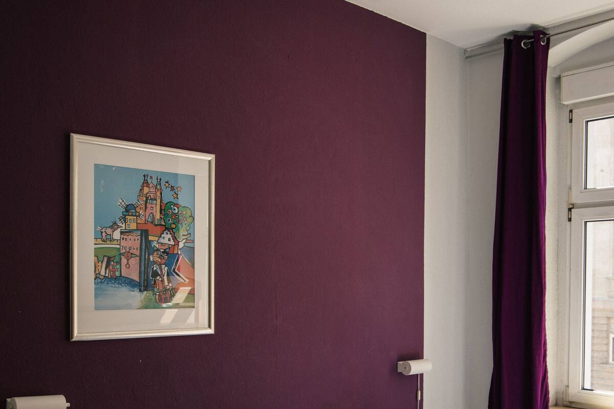 A room with a bed, a nightstand, a framed picture on the wall, and a window with purple curtains.