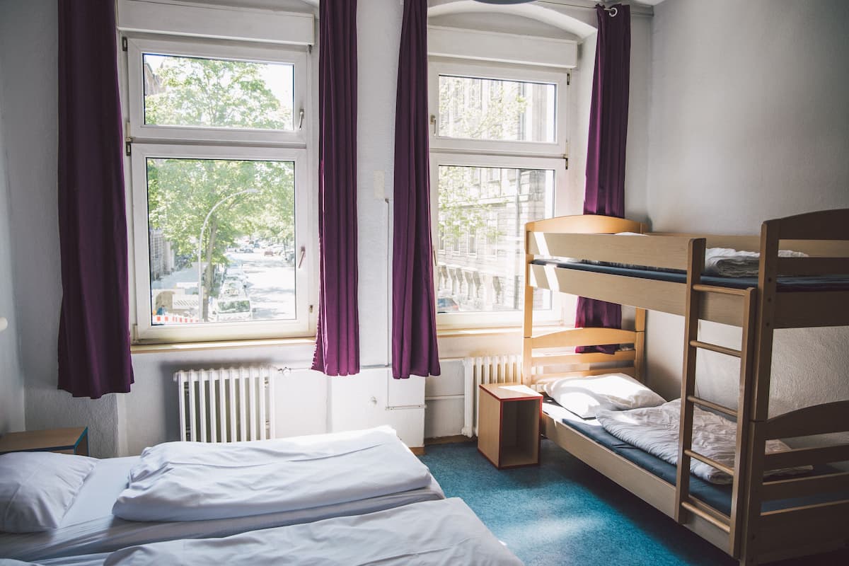 A dormitory room with bunk beds, a window with curtains, a radiator, and a small table.