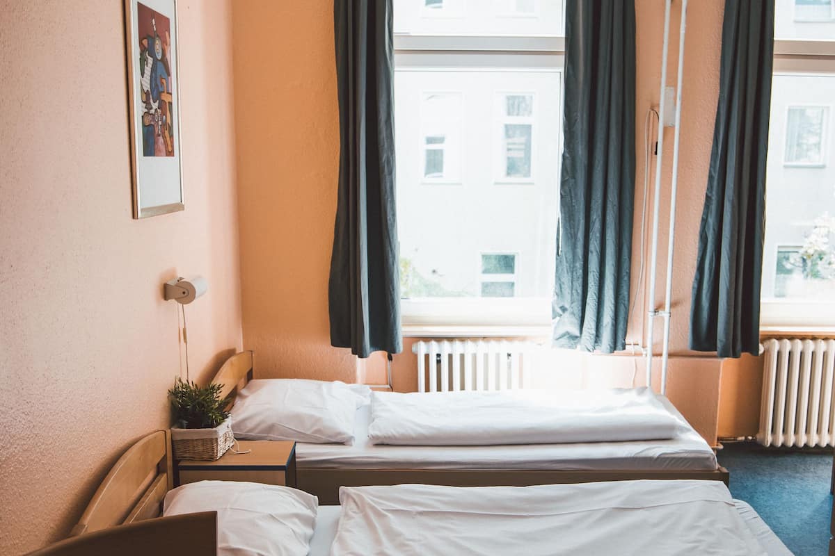 A hotel room with two beds, a desk, a chair, a radiator, and a window with curtains.