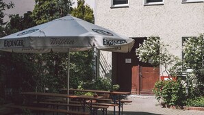 An outdoor seating area with a large umbrella labeled "ERDINGER" and "Weißbier," wooden benches, and a building with multiple windows in the background. - Happy Hotel Berlin (Berlin)
