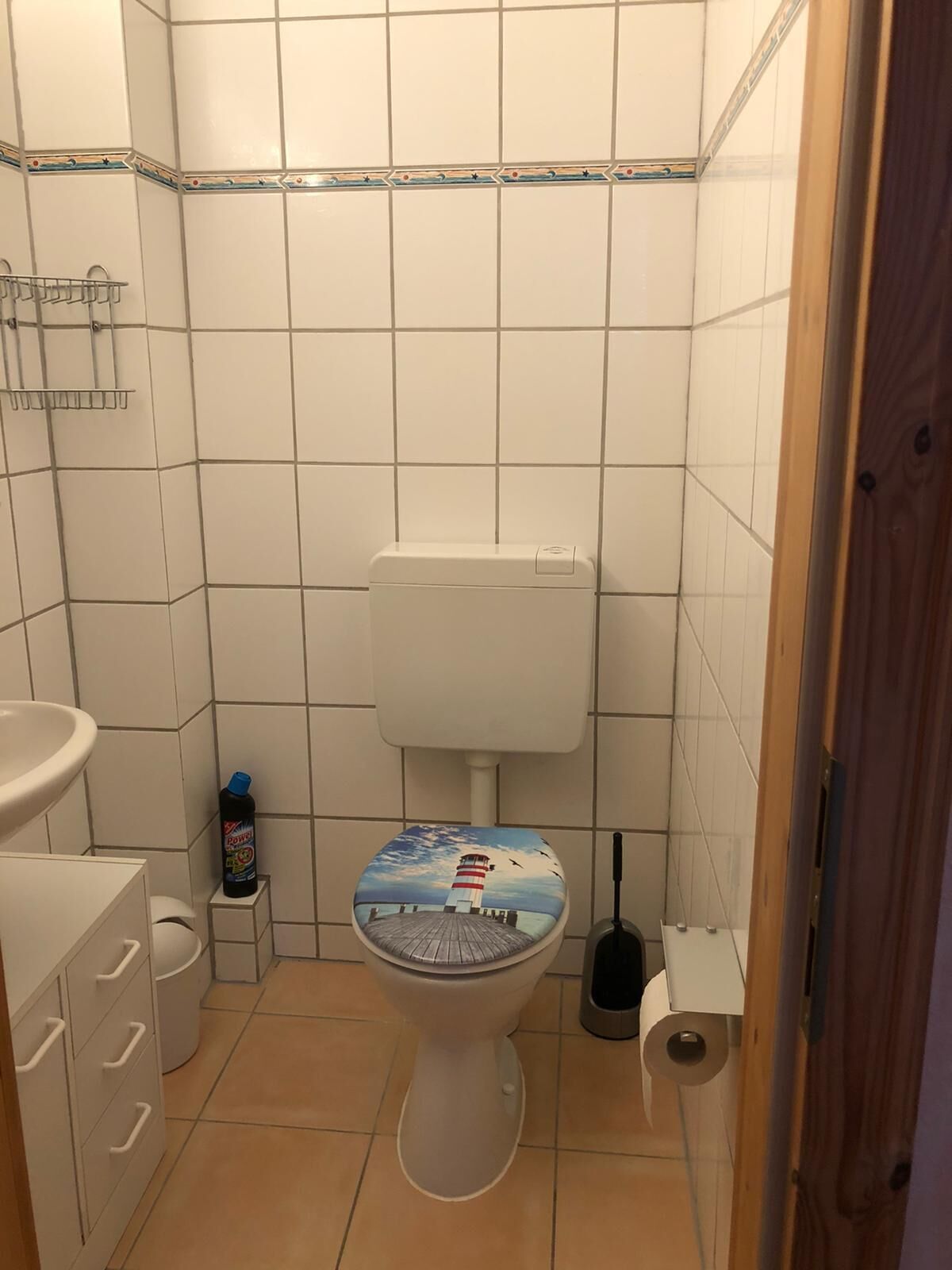 Et lille, rent badeværelse med hvide fliser, et toilet med dekorativt sæde, en vask, en toiletbørste og en flaske rengøringsmiddel.