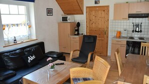 A compact living space with a sofa, a dining table, a kitchen area with wooden cabinets, and a window with curtains. - Vacation home starboard quietly located large terrace only 300m to the beach / sea (Friedrichskoog-Spitze)
