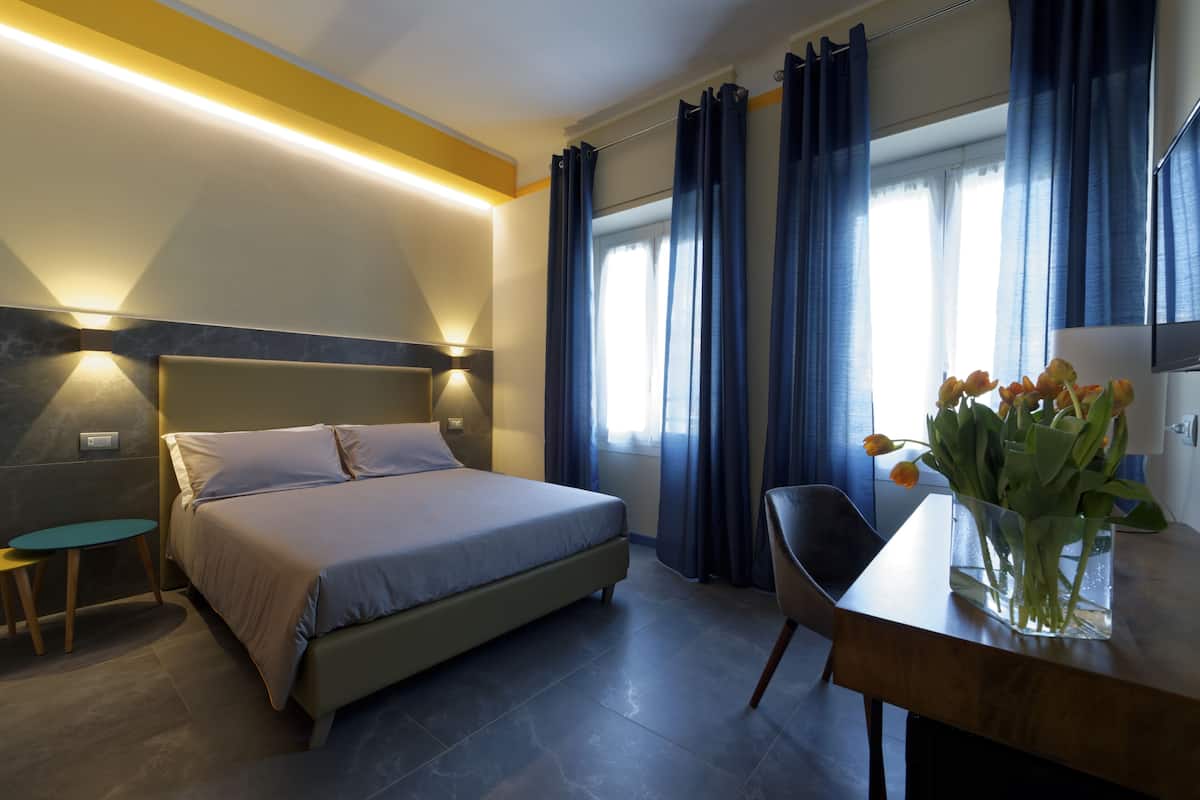 A hotel room with a bed, a desk, a chair, a vase with flowers, and a window with curtains.