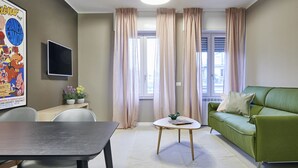 Una sala de estar con un sofá verde, una mesita redonda, un televisor montado en la pared y una ventana con cortinas.