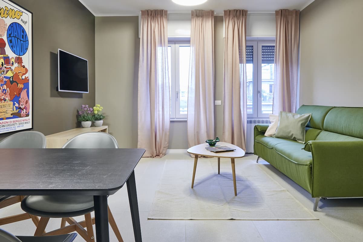 A living room with a green sofa, a small round table, a TV mounted on the wall, and a window with curtains.