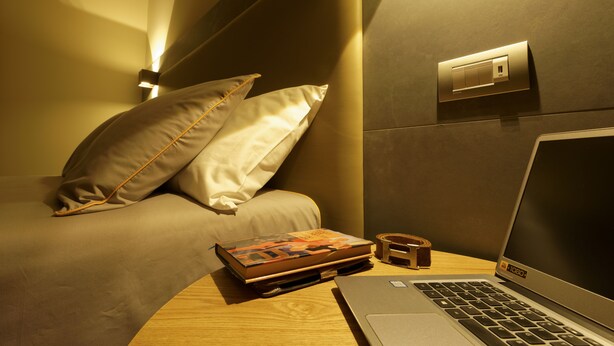 Un dormitorio ordenado con cama, almohadas, una computadora portátil, una mesita de noche con un libro y un pequeño cofre de madera.