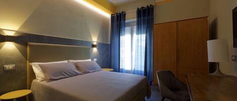 A hotel room with a bed, bedside table, chair, desk, and curtains.