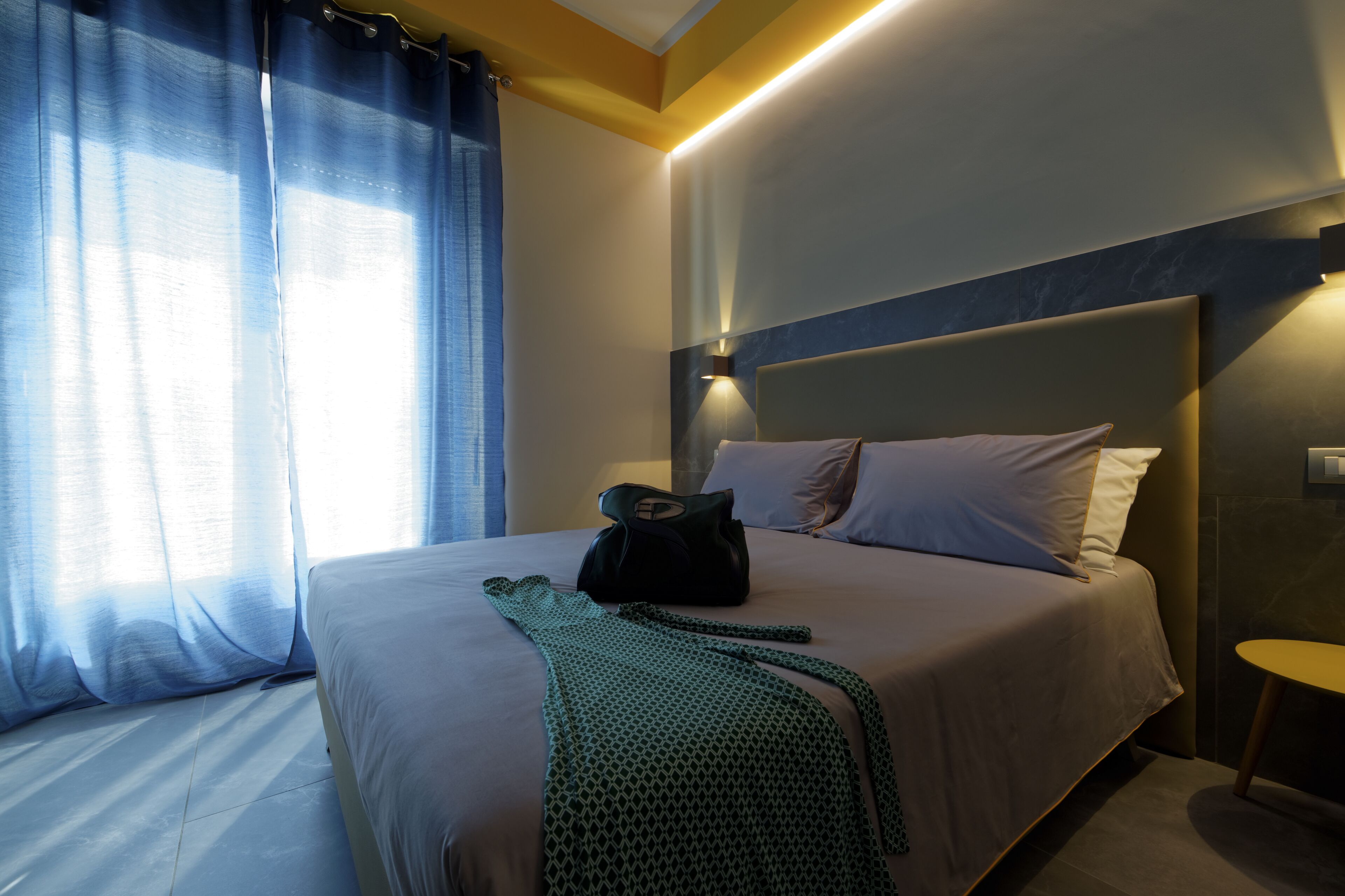 Una habitación de hotel con cama, mesita de noche, lámpara y ventana con cortinas.