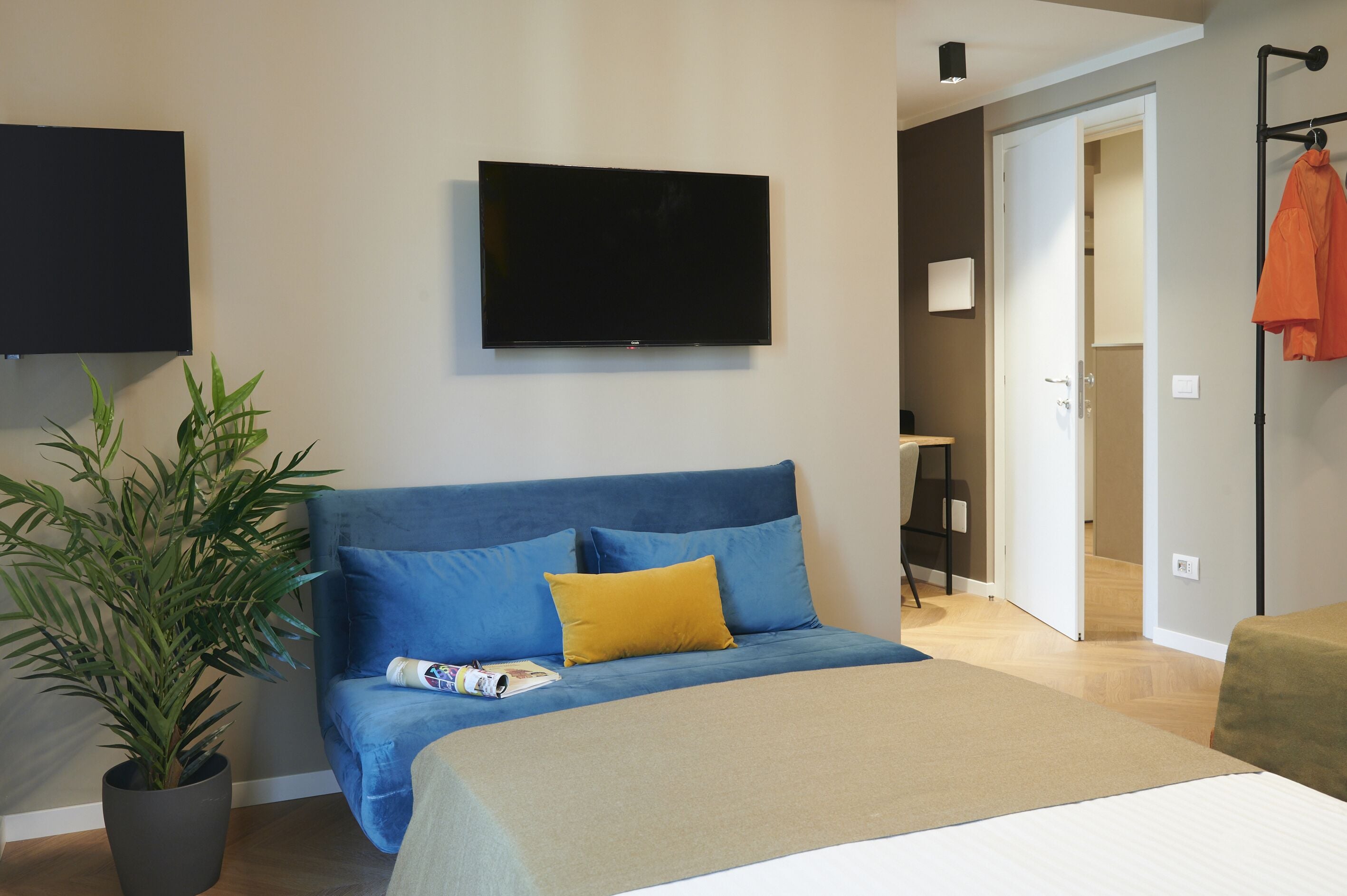 A bedroom with a bed, a television mounted on the wall, a potted plant, and a door leading to another room.