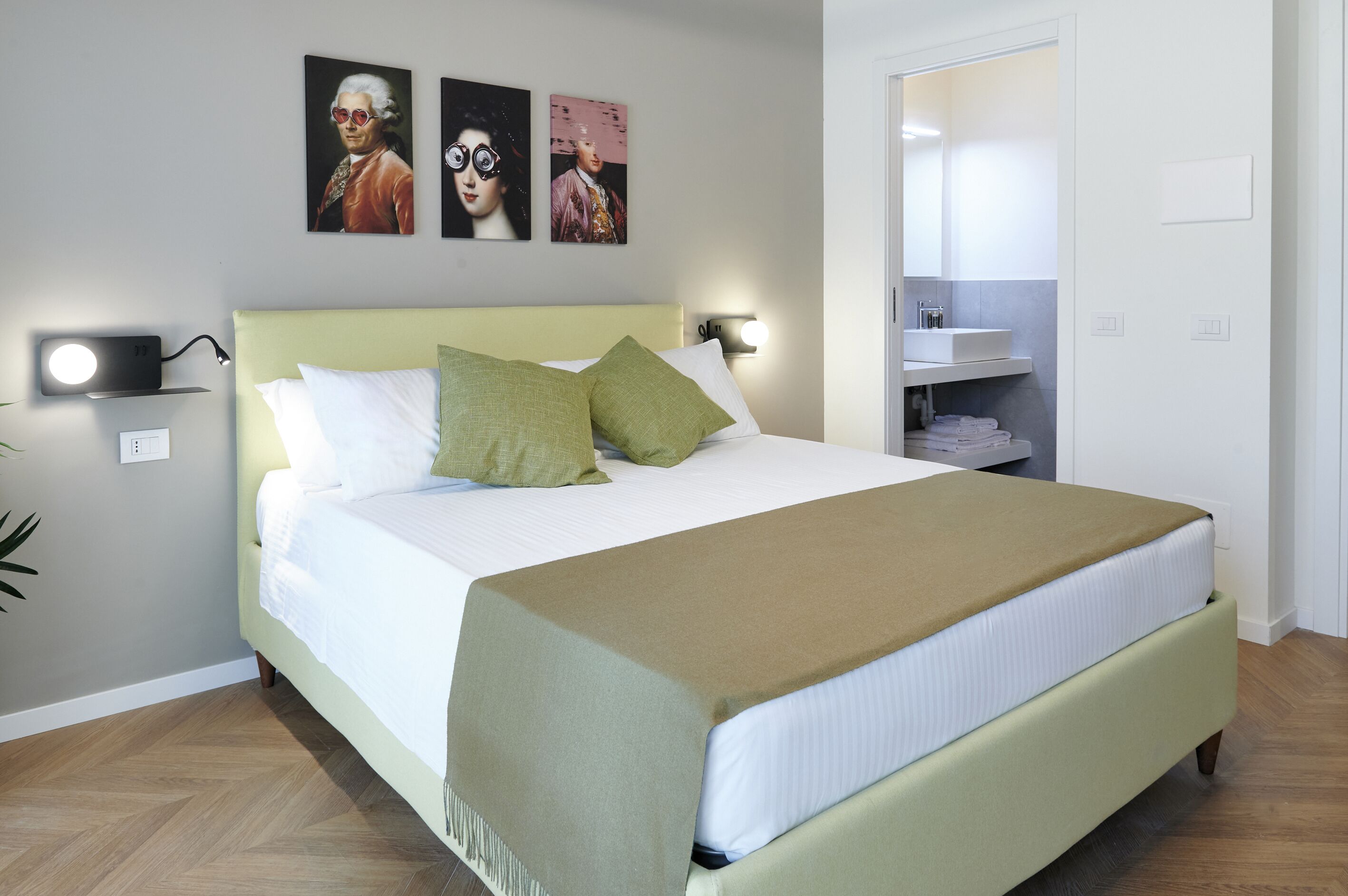 Una cama bien hecha con cabecera verde, ropa de cama blanca y una manta marrón. Hay dos lámparas fijadas en la pared a cada lado de la cama.