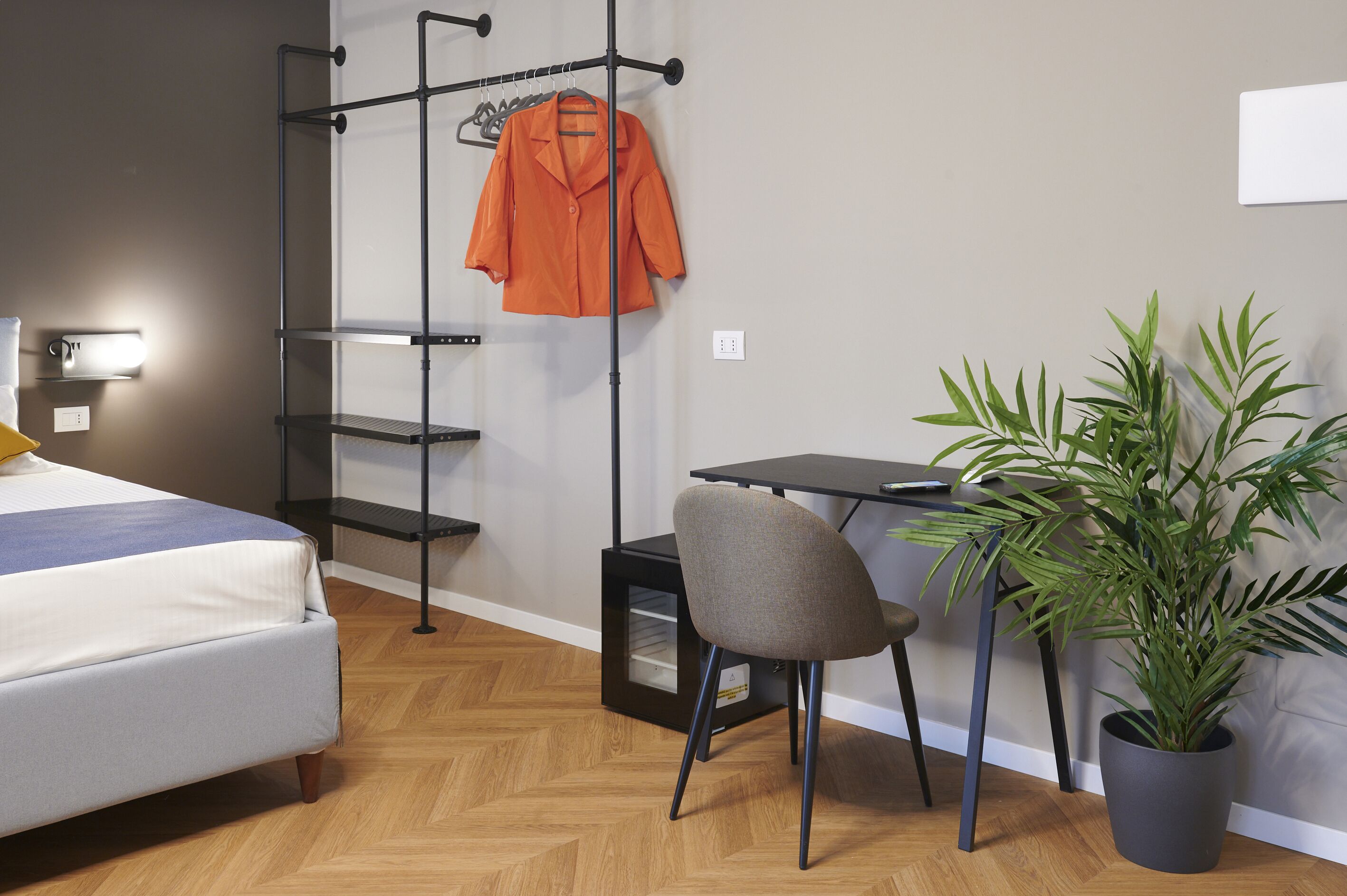 A bedroom with a bed, a desk, a chair, a plant, and a clothing rack.