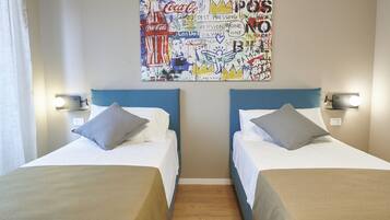 A hotel room with two beds, each with a blue headboard and white bedding. There is a wall art piece above the beds featuring various text and images, including a Coca-Cola bottle.