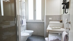 Uma casa de banho moderna com cabine de chuveiro em vidro, lavatório branco, bidé e janela que proporciona luz natural.